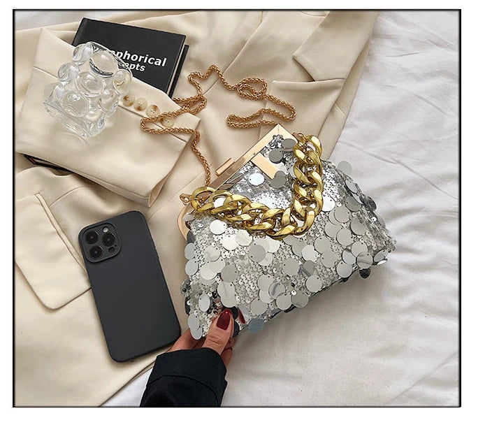 Silver sequin clutch with gold chain held by a hand on a white surface with a beige jacket and phone.